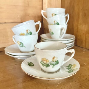 May include: A set of white porcelain teacups and saucers. Each cup and saucer features a delicate floral design with yellow and green accents. The cups have a textured pattern and gold trim, creating an elegant aesthetic. The set includes multiple cups and saucers.