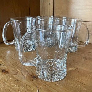 May include: Set of clear glass mugs with handles. The mugs have a textured, bubble-like design on the bottom. The mugs are empty and sitting on a wooden surface.