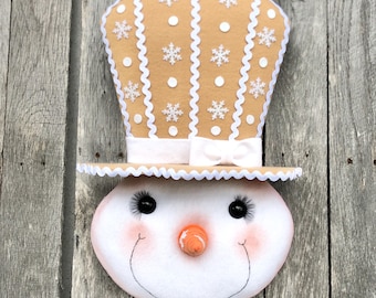 May include: A snowman decoration with a tan top hat adorned with white snowflakes and dots. The snowman's face is white with black eyes, rosy cheeks, a carrot nose, and a stitched smile. The hat has a white bow.