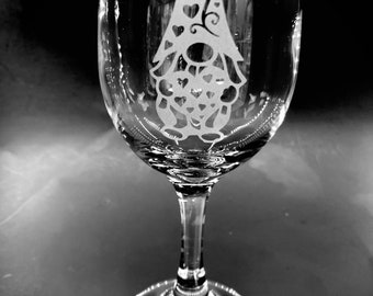 May include: A clear glass wine glass with an etched design of a gnome with a curly hat and a beard. The gnome is surrounded by hearts.