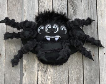 May include: A plush black spider with eight legs and a furry body. The spider has three pairs of white-rimmed eyes and two white fangs. The spider is a Halloween decoration.