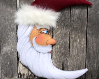 May include: A whimsical crescent moon decoration with a Santa Claus face. The moon is white with a long, flowing beard and a red hat trimmed with white faux fur. The face has rosy cheeks and blue eyes, perfect for holiday decor.