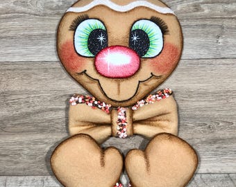 May include: A tan gingerbread-style character cutout featuring a large pink nose, green eyes with long eyelashes, and a white icing-like trim on the head. It includes a tan bow tie and two tan mittens, all accented with small, colorful sprinkles.