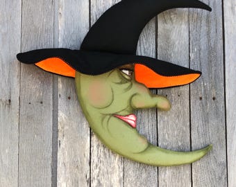 May include: A whimsical Halloween decoration featuring a crescent moon face wearing a black witch hat with an orange brim. The moon has a green complexion, rosy cheeks, and a smiling red mouth. The hat is black with an orange underside.