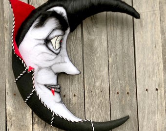 May include: A crescent moon-shaped decorative item with a vampire face. The moon is black with a white face, red accents, and a black wig. The edges are trimmed with black and white rope. The moon has a red drop of blood.
