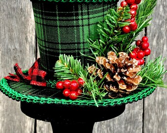 May include: A decorative top hat with a green and black plaid pattern, trimmed with green trim. The hat is adorned with faux pine needles, red berries, and a pine cone. The hat sits on a black pedestal.