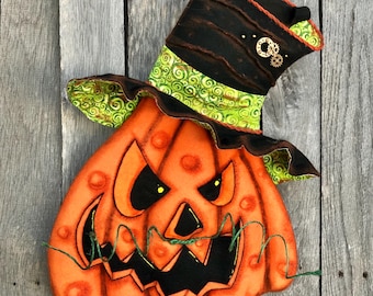 May include: A Halloween decoration featuring an orange pumpkin with a menacing face and a black top hat. The pumpkin has a stitched design with a green and black hat, and yellow eyes. The hat is decorated with gears and orange trim.