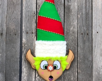 May include: A whimsical elf head decoration with a red and green hat, white fur trim, and a surprised expression. The elf's face is tan with green hair, large eyes, and a snowflake detail. The decoration is mounted on a wooden background.
