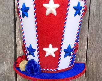 May include: A patriotic Uncle Sam hat with a red, white, and blue color scheme. The hat features white and red sections, adorned with blue and white stars. A blue brim and decorative trim complete the festive design.