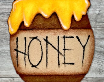 May include: A brown honey pot with a yellow honey drizzle on top. The pot has the word "HONEY" written on the front.