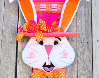 May include: A decorative Easter bunny head with a pink and orange top hat and bow tie. The bunny has large white ears with orange interiors, a pink nose, and a smiling mouth. Small carrot decorations are on the hat.