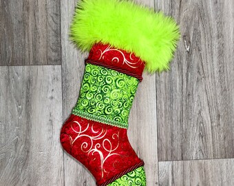 May include: A Christmas stocking with a bright green fluffy top. The stocking is red with gold swirls and green swirl patterns. A red rope trim accents the stocking. The background is a wood grain pattern.