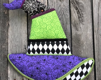 May include: A purple, green, black, and white witch hat with a spiderweb pattern. The hat is made of fabric and has a pointed top.