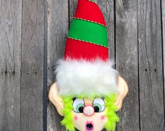 May include: A whimsical elf face decoration with a red and green hat trimmed with gold rope. The elf has a surprised expression, green hair, and a white fluffy trim. The background is a weathered wooden surface.