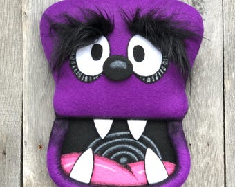 May include: A purple monster-shaped hand puppet featuring large white eyes with black pupils, thick black faux fur eyebrows, a round black nose, and an open mouth with four white pointed teeth and a pink tongue over a black background.