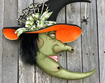 May include: A whimsical Halloween decoration featuring a green crescent moon with a witch-like face. The moon wears a black witch hat with an orange brim, a decorative bow, and a spider dangling from a string. The face has black hair and a pink tongue.