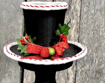 May include: A black top hat with a white and red trim, decorated with a red glitter bow, holly leaves, and red berries. The hat is a festive holiday decoration.