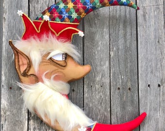 May include: A whimsical crescent moon decoration with a face, elf-like ears, and a red pointed hat. The hat is adorned with gold trim and a colorful star pattern. The moon has a white beard and a red tip.