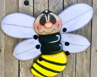 May include: A whimsical wooden bee decoration with a smiling face, white wings, and a black and yellow striped body. The bee has black antennae and is painted with bright colors. This decorative item is perfect for adding a touch of nature to any space.
