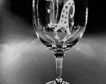 May include: A clear glass wine glass with an etched design of a gnome holding balloons shaped like hearts. The gnome is wearing a hat with hearts on it.
