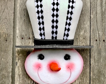 May include: A snowman-shaped wall hanging with a white face and a top hat. The hat is white with black and white diamond-patterned ribbons. The snowman has black eyes, a carrot nose, rosy cheeks, and a smiling mouth. The hat brim is black with white dots.