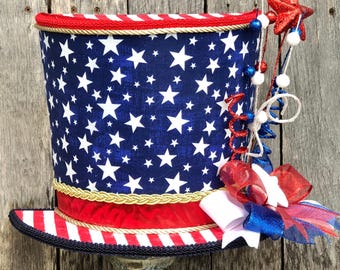 May include: A patriotic top hat with a blue background and white stars. The hat features a red and white striped brim, a red ribbon, and decorative embellishments including a red star and bows. The hat is displayed on a decorative stand.