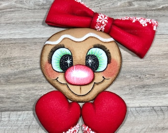 May include: A gingerbread-style face decoration featuring a large red fabric bow with white peppermint accents, green eyes with long black lashes, a round pink nose, and two red heart-shaped mittens with white peppermint trim at the base.