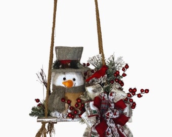 May include: A hanging Christmas decoration featuring two wooden shelves suspended by rope. The lower shelf holds a snowman with a top hat and scarf. The shelves are decorated with red berries, greenery, and a large red and white bow. A red bird sits on the top shelf.