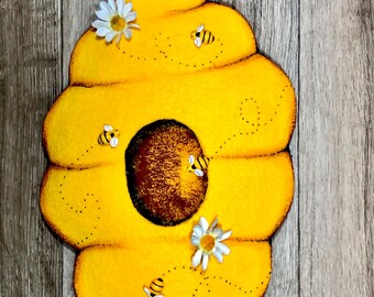 May include: A yellow felt beehive with a brown entrance and four small felt bees flying around it. There are four white felt daisies around the beehive.