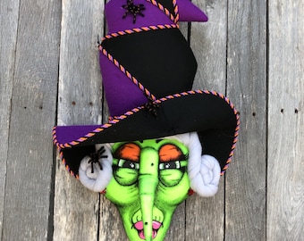 May include: A Halloween-themed witch decoration. The witch has a green face, orange eyes, and a long nose. She wears a black and purple witch hat with orange and purple trim. The hat is adorned with a spider.