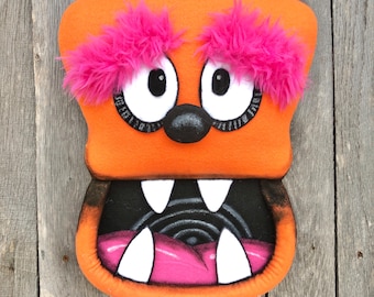 May include: A plush monster character with bright orange fabric, vibrant pink faux fur eyebrows, and large white eyes with black pupils. The creature features a wide open mouth with white felt teeth, a pink tongue, and a black nose.