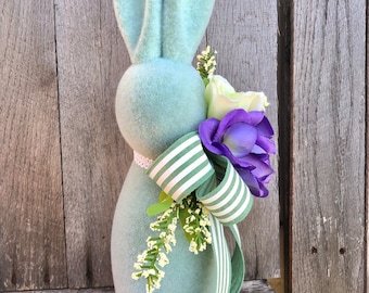 May include: A light blue plush bunny rabbit with a white and green striped ribbon and purple and white flowers.