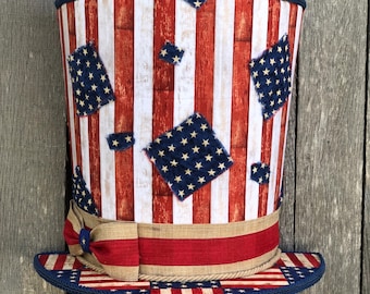May include: A patriotic top hat with a red, white, and blue design. The hat features vertical red and white stripes, blue star-studded patches, and a red and tan ribbon. The brim and trim are navy blue.