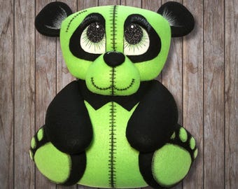 May include: A plush panda bear with a lime green body and black accents. The bear has large, glittery eyes, a stitched design, and a friendly smile. The ears, paws, and arms are black. The background is a wooden surface.