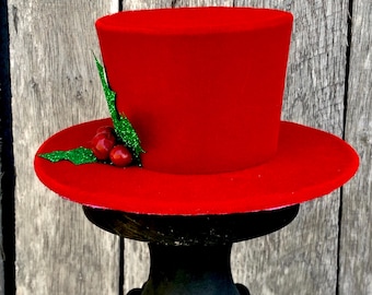 May include: A bright red top hat with a wide brim, decorated with green glitter leaves and red berry accents. The hat is displayed on a black pedestal, perfect for holiday or festive occasions.