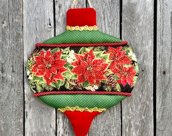 May include: A decorative Christmas ornament in an elongated teardrop shape. The ornament features a red velvet top and bottom, with a green polka-dot center. The center panel displays red poinsettias and green leaves. Gold trim and red rope detail the edges.