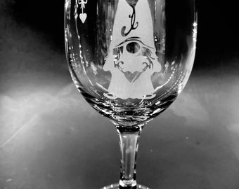 May include: A clear glass wine glass with an etched design of a gnome wearing a hat with a heart on it.
