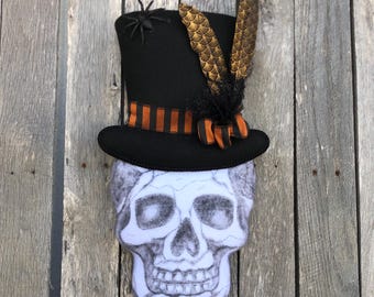 May include: A Halloween-themed decoration featuring a skull with a black top hat. The hat is adorned with an orange and black striped ribbon, a small black spider, and decorative feathers. The skull is white with gray shading.