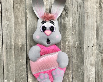 May include: A gray felt bunny with pink ears and a pink dress. The bunny has large, black eyes and a pink nose. The bunny is holding a pink egg with a white lace trim.