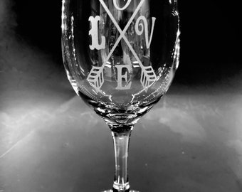 May include: A clear glass wine glass with an etched design of two arrows crossing with the word "LOVE" and a heart in the center.