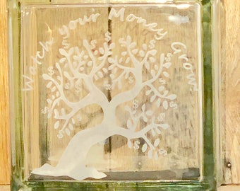 May include: A clear glass block with a tree design etched on the front. The tree has dollar signs on its leaves, and the words "Watch your Money Grow" are etched around the tree.