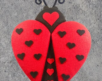 May include: Red felt ladybug with black felt accents and a heart design. The ladybug has black felt antennae and a large heart on its back. The ladybug is covered in small black felt hearts.