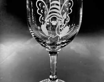 May include: A clear wine glass with a whimsical gnome design etched on the front. The gnome has a striped hat, wings, and is surrounded by hearts and crowns. The glass has a long stem and a round base.