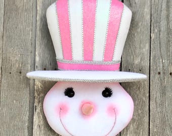 May include: A snowman face decoration with a white top hat featuring pink stripes and silver trim. The snowman has black eyes, a pink nose, rosy cheeks, and a smiling mouth. The background is weathered wood.