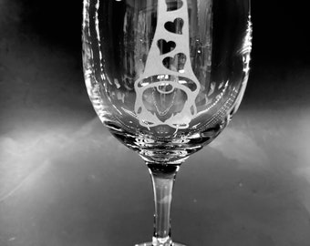 May include: A clear wine glass with a decorative gnome design etched into the glass. The gnome's hat is adorned with heart shapes. The glass has a long stem and a round base, suitable for serving wine.