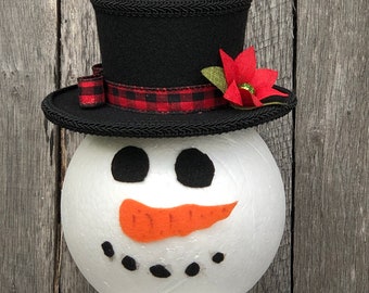 May include: A snowman head with a black top hat adorned with a red and black plaid ribbon and a red poinsettia. The snowman has black button eyes, an orange carrot nose, and black coal mouth dots. The snowman is a festive holiday decoration.