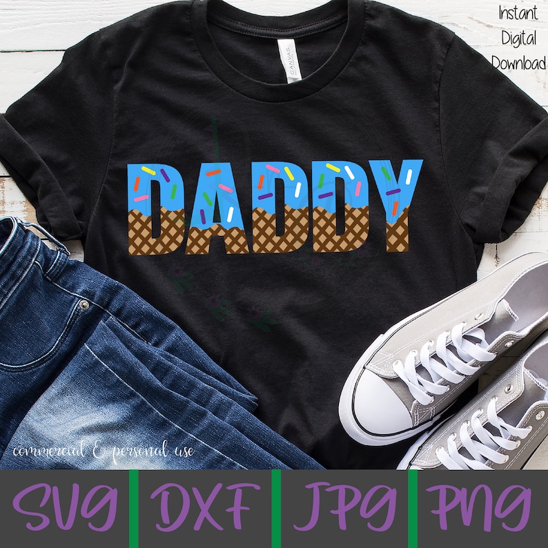 Puede incluir: Una camiseta negra con la palabra "DADDY" en letras azules con un cono de waffle marr&oacute;n y chispas de colores en la parte superior. La camiseta est&aacute; sobre una superficie blanca con un par de zapatillas grises y jeans azules.