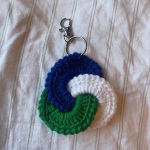 May include: A crocheted keychain with a blue, white, and green swirl design. The keychain is attached to a silver metal ring and a silver metal clasp.