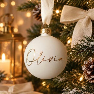 May include: A matte white spherical Christmas ornament featuring the name Olivia written in elegant gold cursive script. The ornament is topped with a gold metal cap and hangs from a cream-colored satin ribbon.