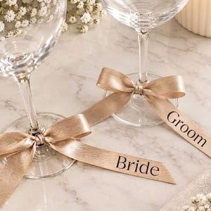 May include: Two clear glass wine glasses, each featuring a champagne-coloured satin ribbon tied around the stem. One ribbon is printed with the word Bride in black, and the other ribbon is printed with the word Groom in black lettering.